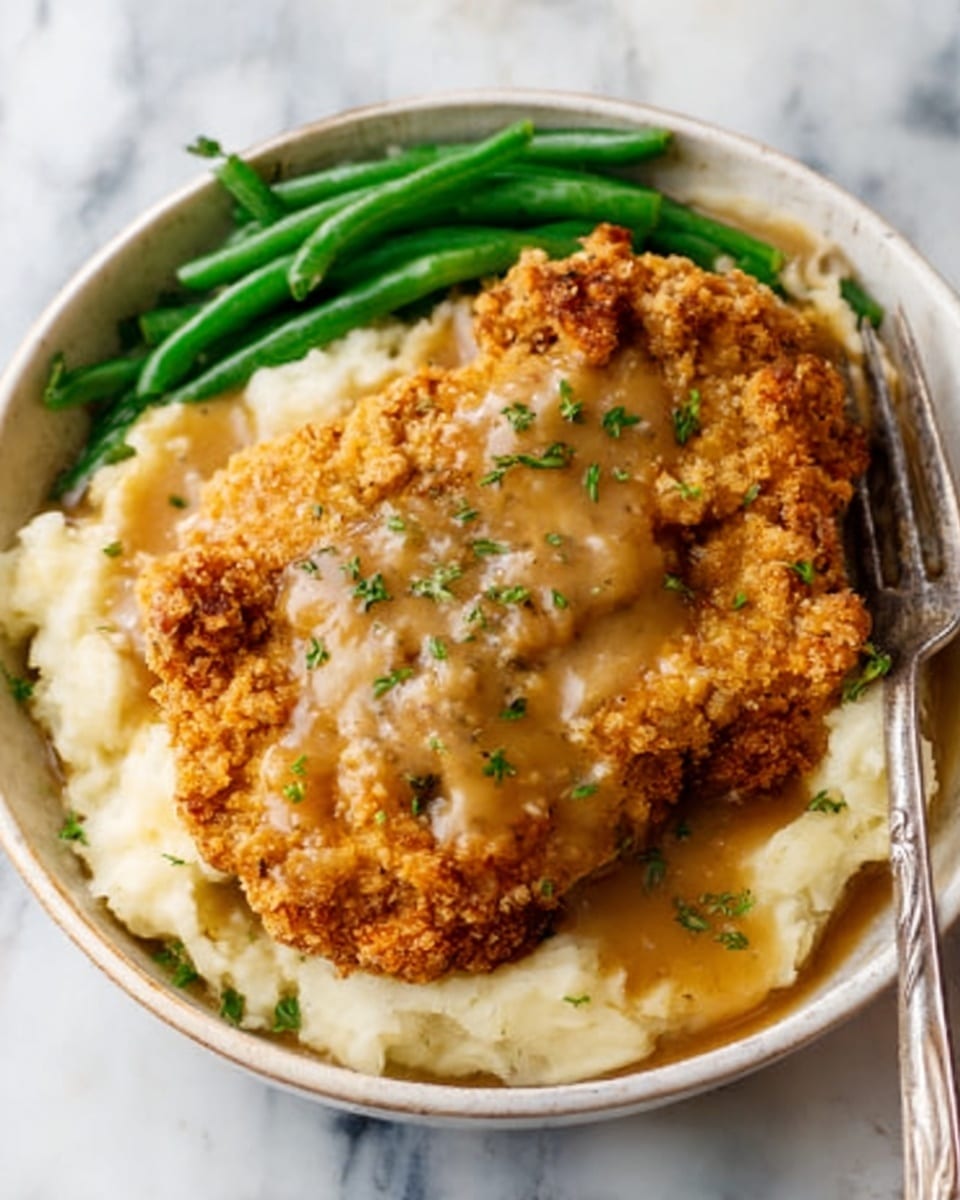 A white bowl filled with creamy mashed potatoes topped with golden brown chicken fried steak covered in light brown gravy, placed beside a small portion of bright green cooked green beans. The chicken fried steak layer is crispy and rough textured, sitting on top of smooth, thick mashed potatoes with a shiny gravy layer flowing over the steak and potatoes. A silver fork is partially visible inside the bowl, on a white marbled surface. Photo taken with an iphone --ar 4:5 --v 7
