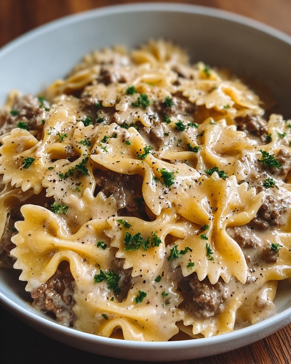 The image shows a close-up of a white bowl filled with creamy pasta made of farfalle (bow-tie) noodles. The first layer is the cooked pasta with a light golden color, covered evenly with creamy beige sauce that has a smooth texture. Mixed in are small brown ground meat pieces scattered throughout the dish. On top, there are small fresh green parsley bits sprinkled across the pasta. The entire dish is lightly dotted with black pepper, adding contrast. The bowl sits on a wooden surface with soft natural light highlighting the creamy texture and fresh herbs on the dish. Photo taken with an iphone --ar 4:5 --v 7