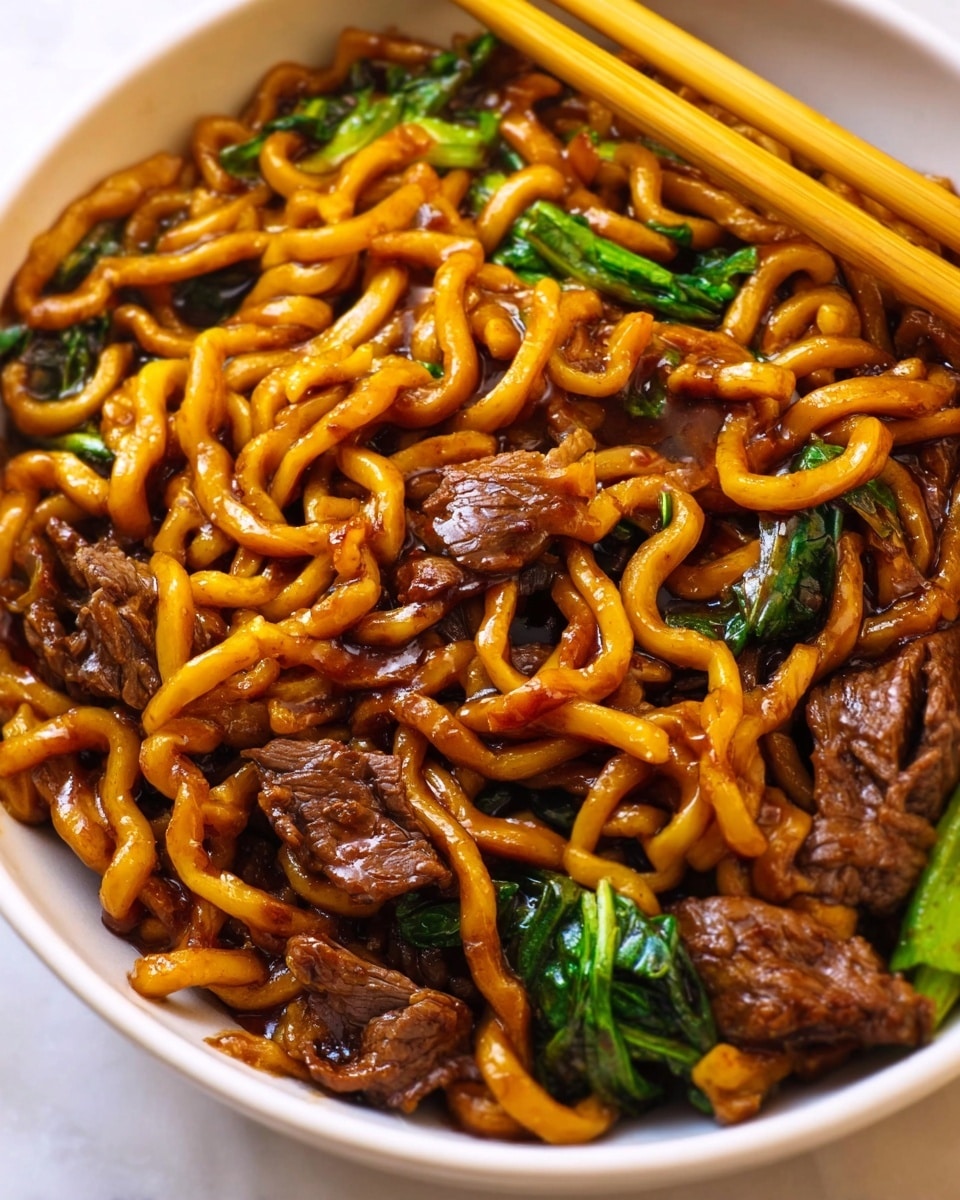 A white bowl filled with thick, glossy udon noodles coated in a rich, dark brown sauce, mixed with tender slices of beef that have a slightly caramelized texture. Bright green leafy vegetables add a fresh pop of color scattered on top and around the meat. Light wooden chopsticks rest on the edge of the bowl, which sits on a white marbled surface. Photo taken with an iphone --ar 4:5 --v 7