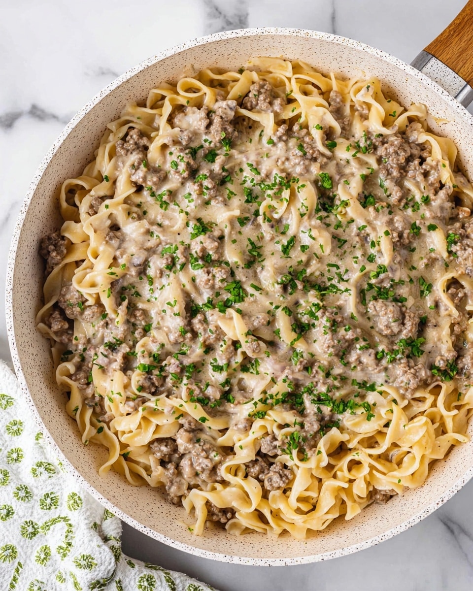 A creamy pasta dish shown in a white pan with speckles, filled with three main layers. The bottom layer consists of light beige egg noodles with a curly and soft texture, spread all across the pan. On top of the noodles is a layer of ground meat in small, chunky pieces coated in a light beige creamy sauce that has a smooth, glossy texture. The dish is finished with a sprinkle of bright green chopped herbs scattered evenly over the top, adding a fresh contrast to the creamy and meaty elements. The pan is placed on a white marbled surface, with a white and green patterned cloth partially visible on the side. Photo taken with an iphone --ar 4:5 --v 7