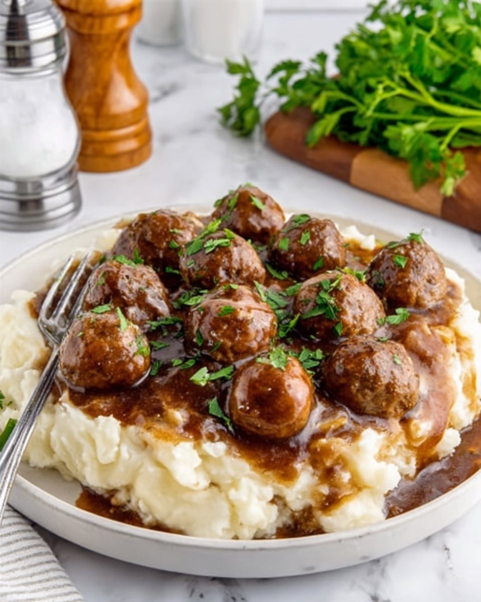 A white plate filled with a thick layer of creamy mashed potatoes, topped with a rich, dark brown gravy that slightly seeps into the potatoes. On top of the gravy, there are several round, browned meatballs garnished with small pieces of fresh green parsley. In the front left of the plate, a stainless steel fork rests partially on the mashed potatoes. The background has a white marbled texture, with a salt shaker, a pepper shaker, and a bunch of fresh green parsley in a wooden container visible but out of focus. Photo taken with an iphone --ar 4:5 --v 7