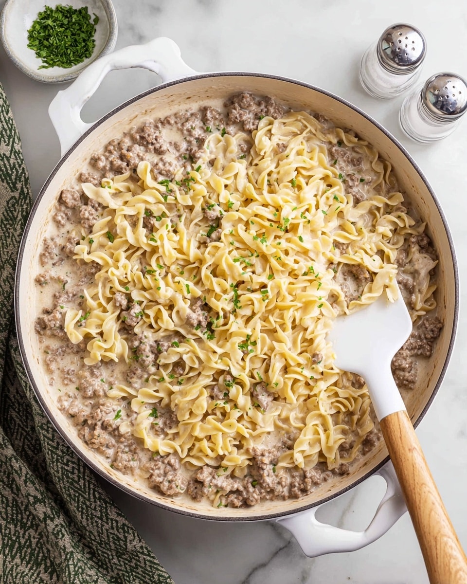A large white pan filled with a creamy pasta dish made of three visible layers: the bottom layer is a sauce with a light beige color and smooth texture, the middle layer is cooked ground meat with a darker brown, crumbly texture evenly mixed throughout, and the top layer consists of soft yellow egg noodles covered lightly in sauce and garnished with small green chopped herbs. A white spatula with a wooden handle is partially under the noodles on the right side of the pan, resting on a white marbled surface with a salt shaker and a green patterned cloth napkin nearby. Photo taken with an iphone --ar 4:5 --v 7