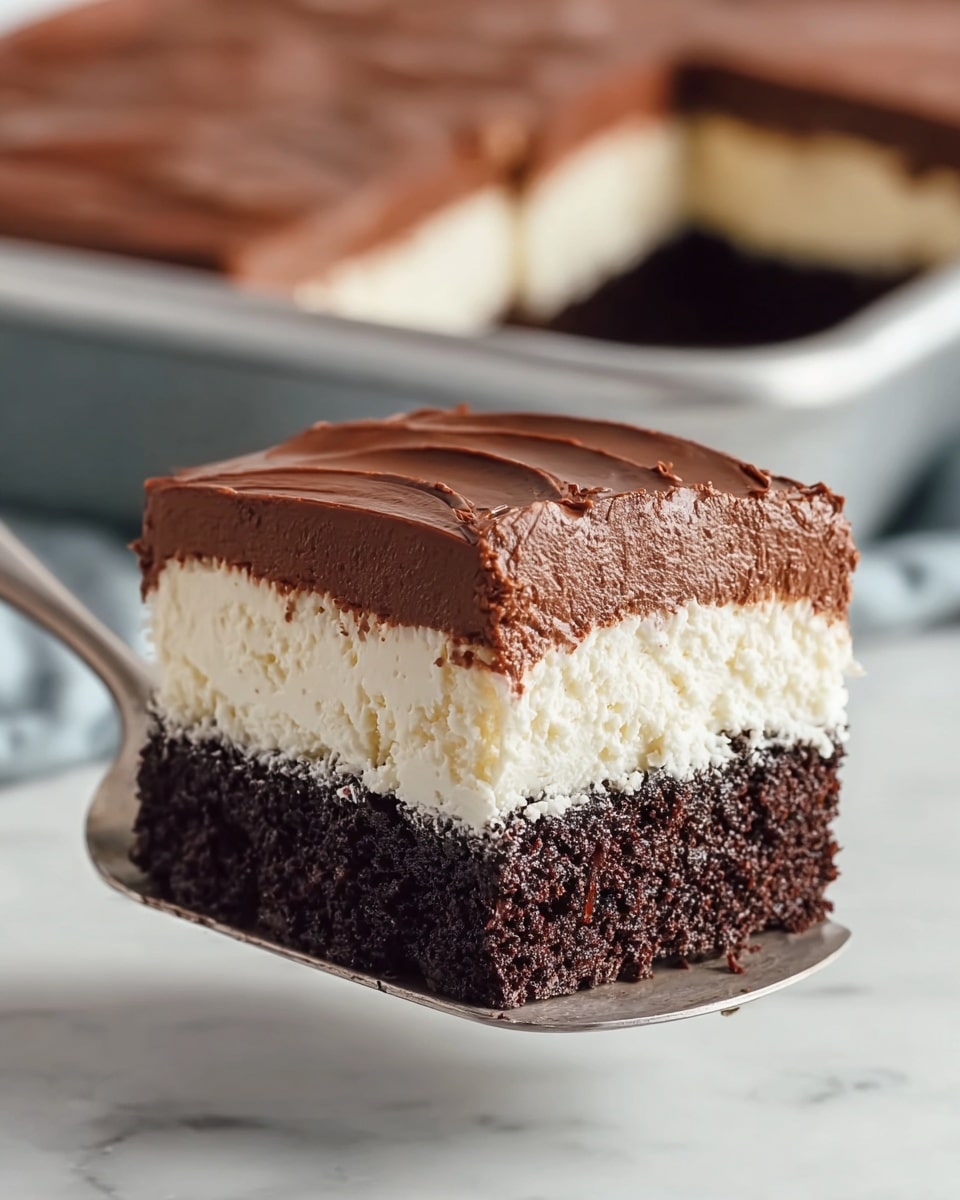 A close-up view of a three-layered dessert square held on a silver utensil, showing a bottom layer of moist dark brown chocolate cake with a slightly crumbly texture, a middle layer of thick, creamy white filling with a smooth, sponge-like surface, and a top layer of velvety chocolate frosting with soft peaks and a shiny finish, with a larger tray of the same dessert visible blurred in the background on a white marbled surface. photo taken with an iphone --ar 4:5 --v 7