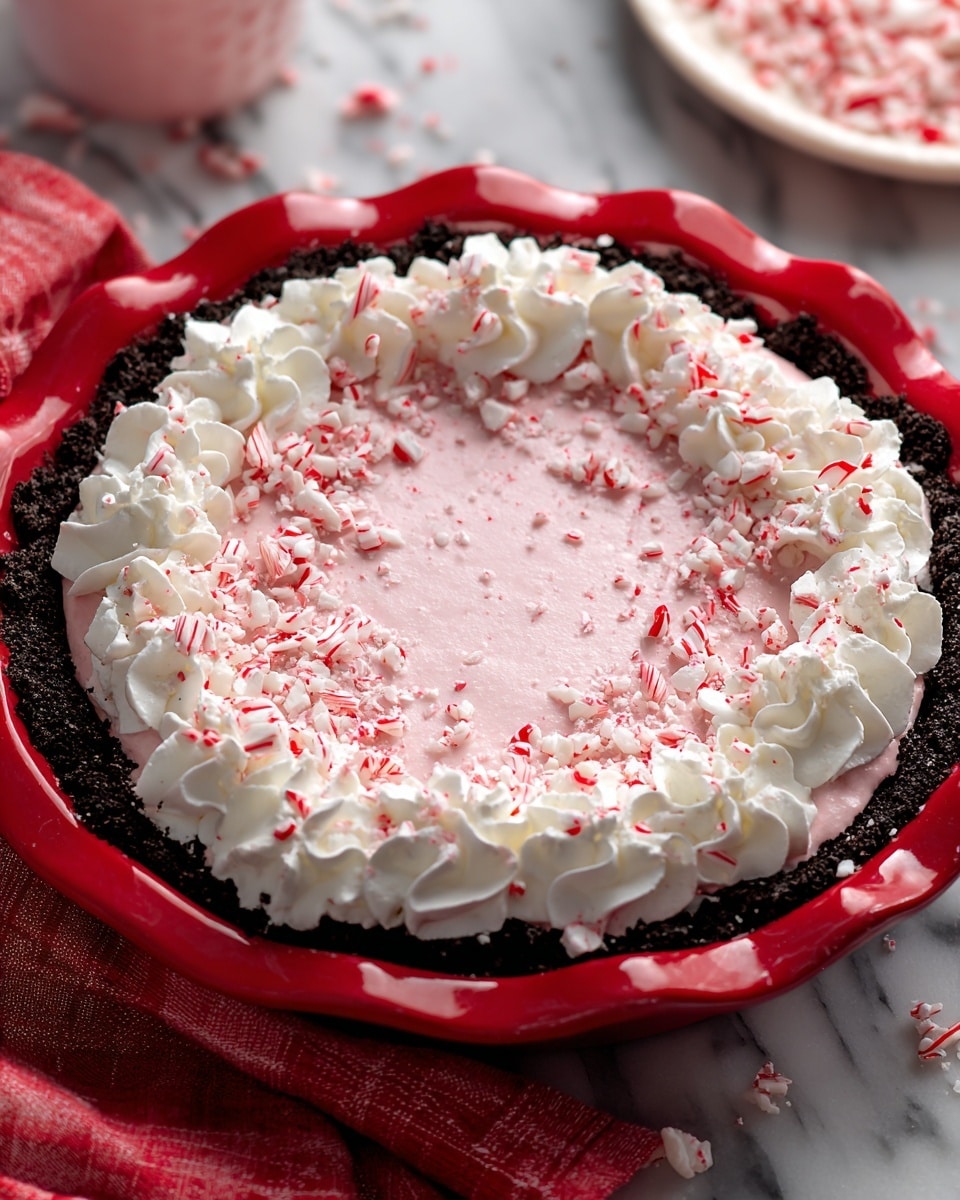 The image shows a three-layer pie in a red pie dish on a white marbled surface. The bottom layer is a dark chocolate crumb crust that edges the dish. The middle layer is a smooth, light pink creamy filling that fills the pie. The top layer has white whipped cream piped in dollops around the edge, with crushed red and white peppermint candy sprinkled all over the pink cream and whipped cream. The colors are festive with reds, whites, and dark browns, and the scene has a holiday feel. photo taken with an iphone --ar 4:5 --v 7