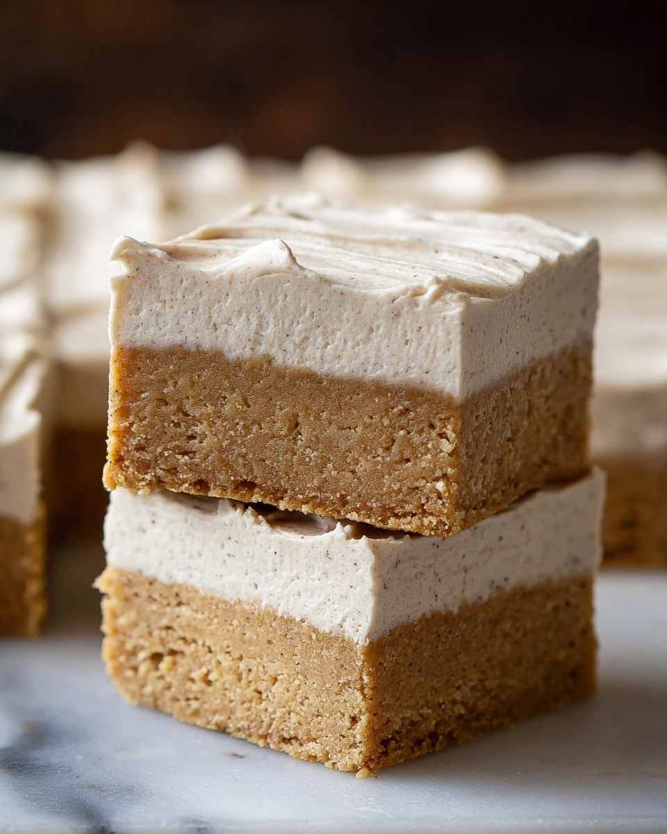 The image shows two dessert bars stacked on top of each other, each bar having two clear layers. The bottom layer is thick, dense, and golden brown with a slightly moist texture that looks soft and cake-like. The top layer is a thick, creamy off-white frosting with small flecks, giving it a lightly textured and whipped appearance with soft peaks on the edges. Additional bars of the same dessert can be seen blurred in the background, resting on a white marbled surface. Photo taken with an iphone --ar 4:5 --v 7