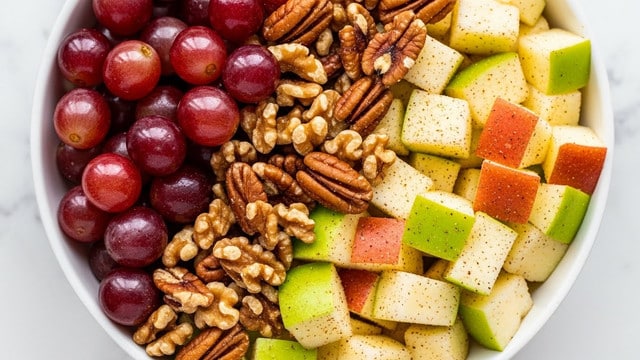 The image shows a close-up of a fruit and nut salad in a white bowl on a white marbled surface. The salad has three main layers: shiny red grapes scattered evenly, pale yellow and green apple cubes with a fuzzy texture, and crunchy brown pecans and walnuts mixed throughout. The fruit and nuts are coated lightly with a dusting of spice or seasoning, adding tiny specks of dark brown on their surfaces. The photo captures the fresh, juicy look of the fruit and the rich, textured shells of the nuts. photo taken with an iphone --ar 4:5 --v 7