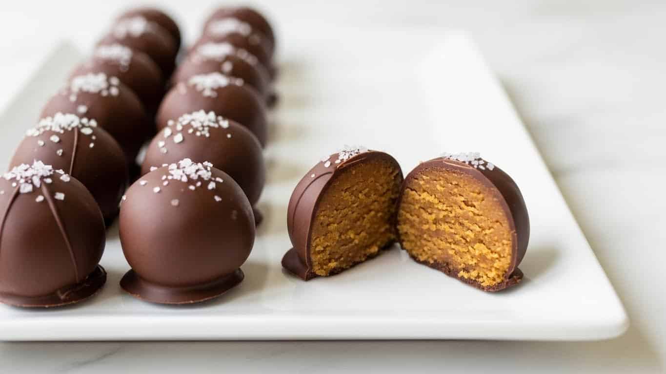 A white rectangular plate holds two rows of round chocolate truffles, each coated in a smooth dark brown chocolate shell. Each truffle is topped with a small sprinkle of coarse white salt. The truffle in the middle front is cut in half, showing a dense, slightly crumbly golden brown inside. The plate sits on a white marbled surface, and the image is taken from a low angle right in front of the truffles, making the details of the texture on the cut piece stand out clearly. Photo taken with an iphone --ar 4:5 --v 7