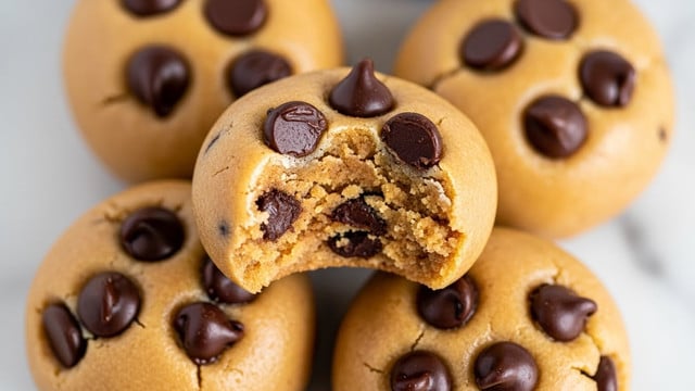 The image shows a close-up of round peanut butter cookie dough balls mixed with dark chocolate chips. The top layer is smooth and light brown with several dark chocolate chips scattered on the surface, some slightly melted. One cookie dough ball in the center has a bite taken out, revealing a dense, slightly grainy inside with some chocolate chips embedded throughout. The cookie dough balls are stacked closely together on a white marbled surface. photo taken with an iphone --ar 4:5 --v 7