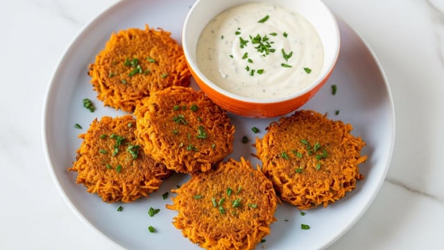 The image shows several thick, round, orange patties cooking in a skillet filled with oil, each with a crispy, dark brown crust and a textured surface made up of small grains. Small green herb leaves are sprinkled on top of and around the patties, adding a pop of fresh color. The patties are layered with a slightly rough texture indicating grains or vegetables inside, and the oil glistens around them, making them look juicy and well-fried. The skillet is black, and everything is set against a white marbled texture. photo taken with an iphone --ar 4:5 --v 7