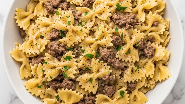 A close-up view of a white bowl filled with farfalle pasta mixed with ground beef in a creamy sauce, sprinkled evenly with small green parsley pieces and black pepper dots. The pasta is yellowish with a smooth, saucy texture, and the ground beef appears brown and crumbly, spread throughout the bowl. The dish looks warm and rich, placed on a white marbled surface. photo taken with an iphone --ar 4:5 --v 7