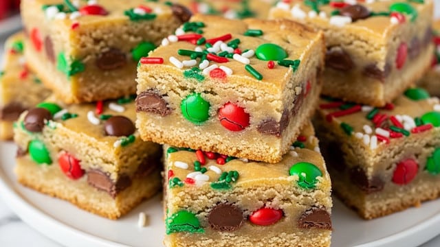 The image shows a close-up of several square pieces of cookie bars stacked in a pile. Each bar has three visible layers: a thick, soft light golden brown base with a slightly crumbly texture, a middle layer filled with colorful candy pieces including red, green, and brown chocolate buttons, and a top layer that looks slightly crisp and golden with white, red, and green sprinkle decorations spread unevenly on the surface. The pieces are set on a white plate partly visible, with a white marbled surface underneath. Photo taken with an iphone --ar 4:5 --v 7