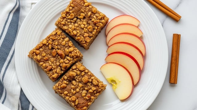 On a white plate with subtle circular ridges, there are three square pieces of crumbly apple nut cake placed closely together on the left side. The cake shows a golden-brown baked crust with visible nut pieces embedded throughout its rough texture. On the right side of the plate, five thin slices of fresh apple with red edges and pale yellow interiors are fanned out neatly. The plate rests on a white marbled surface, accompanied by two cinnamon sticks and a blue-and-white striped cloth partially visible at the bottom left. Photo taken with an iphone --ar 4:5 --v 7