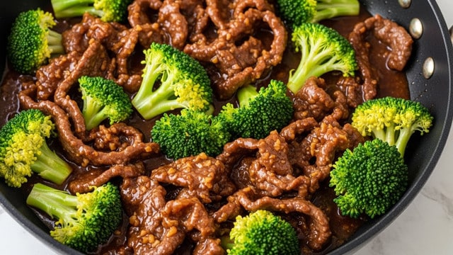 This image shows a close-up of a stir fry in a black pan. The dish has two main layers: tender, thin slices of cooked beef that are deep brown and glossy with sauce, and bright green broccoli florets scattered evenly throughout. The beef pieces are slightly curled and covered in a thick, dark brown sauce with small bits of garlic or ginger visible. The broccoli adds a fresh, textured contrast with its bumpy surface and lighter green stems. The scene sits on a white marbled texture. photo taken with an iphone --ar 4:5 --v 7