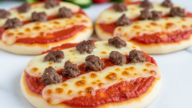 The image shows three small round pizzas placed on a white marbled surface. Each pizza has three visible layers: a light beige crust base, a smooth red tomato sauce next, and a top layer of melted golden brown cheese with small browned spots. There are scattered pieces of cooked ground meat, dark brown in color, evenly spread on the cheese layer. The pizzas are arranged close together in the foreground with a blurred background that seems to include a salad with green and red vegetables. The overall lighting is bright and natural, highlighting the melted cheese texture. Photo taken with an iphone --ar 4:5 --v 7